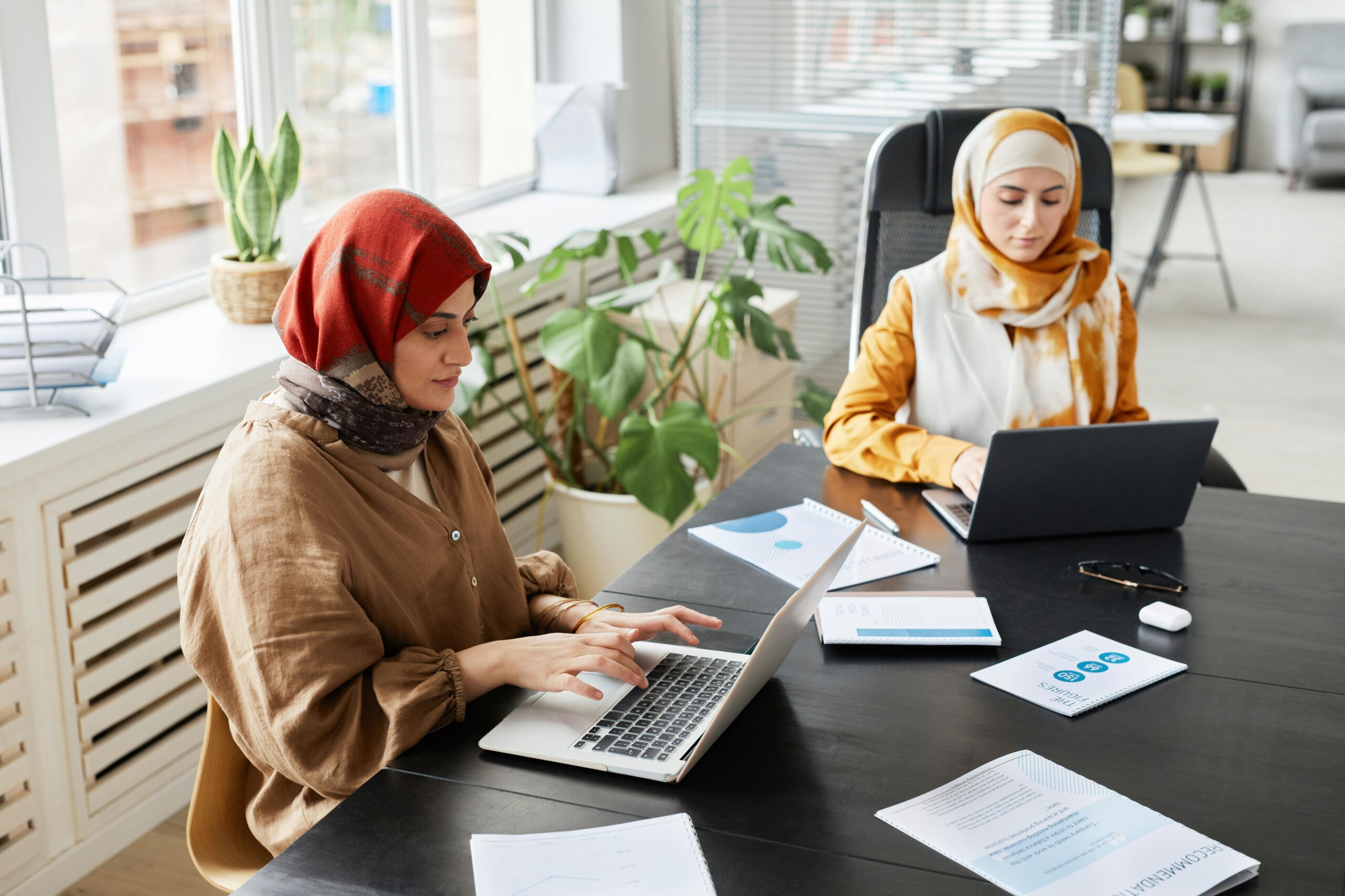 Two Muslim women collaborate using laptops in a bright, plant-filled office.